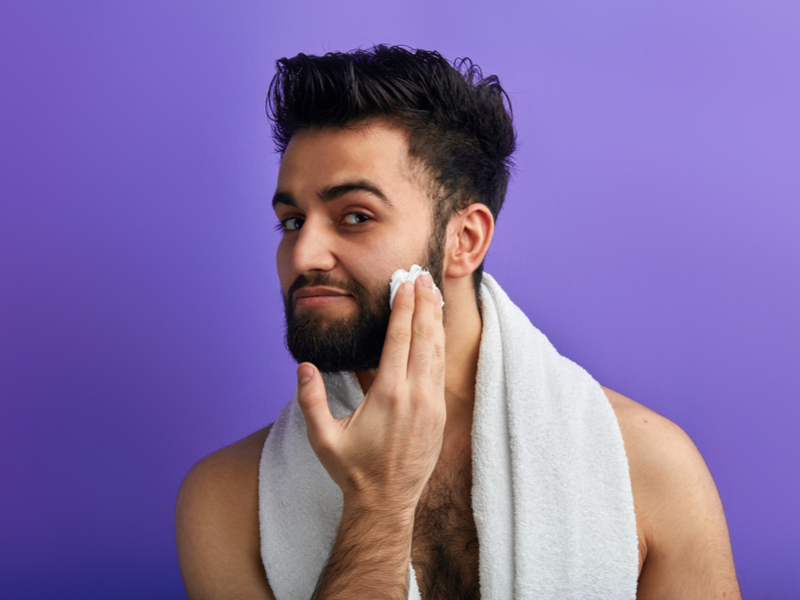 É fundamental cuidar da barba após a coloração Homem cuidando da barba com uma toalha enrolada no pescoço