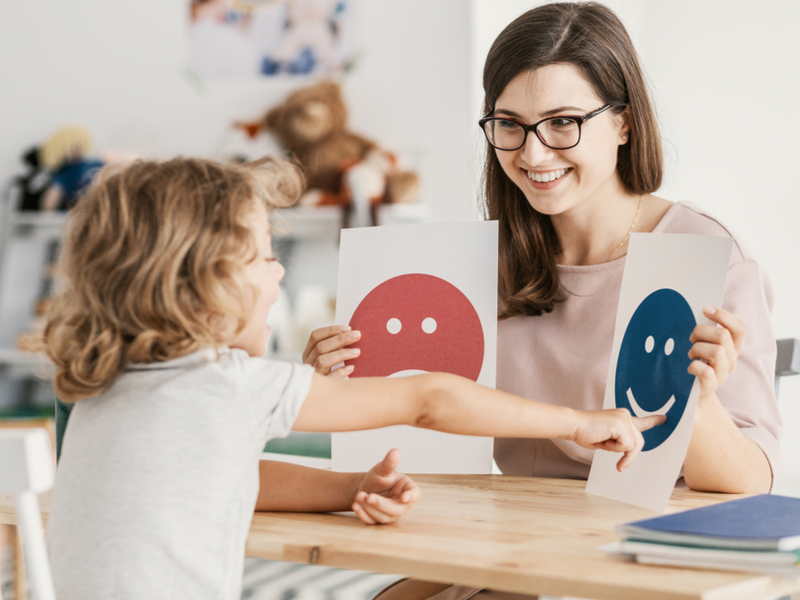 Tratamento para autismo é realizado de forma multidisciplinar Mulher segurando dois papéis com emoji feliz e triste e uma criança apontando para o papel